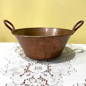 May include: A hammered copper bowl with two arched handles. The bowl has a wide, shallow shape and a rich, reddish-brown color. The handles are a darker shade of brown. The bowl sits on a white lace tablecloth.