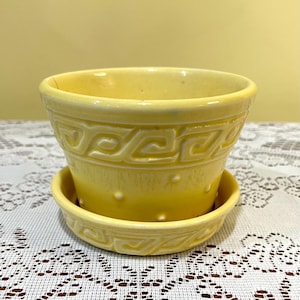 May include: A bright yellow ceramic plant pot with a matching saucer. The exterior features a raised Greek key pattern around the upper rim and a series of small, evenly spaced raised dots encircling the lower body of the cylindrical vessel.