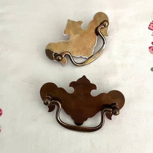 May include: Two antique brass drawer pulls with ornate designs. Each pull features a curved handle and decorative backplate. The brass has a warm, aged patina, suggesting vintage or repurposed use. The pulls are likely for furniture restoration.