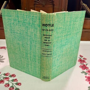 May include: A hardcover book titled Hoyle Up-To-Date Official Rules for All Important Games by Morehead and Mott-Smith, published by Grosset and Dunlap. The book features a textured, light green cover with black text printed on the spine.