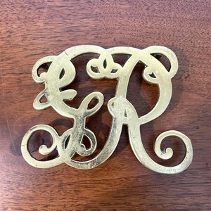 May include: A gold-colored metal monogram letter "R" with a decorative, cursive design. The letter is displayed on a dark brown wooden surface. The letter has a slightly aged appearance.