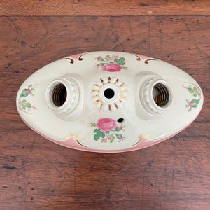 Puede incluir: Luminaria ovalada vintage con dos portalámparas. De color crema, con detalles en rosa y dorado. La lámpara presenta diseños florales de rosas y hojas. Probablemente hecha de cerámica, tiene un diseño decorativo.