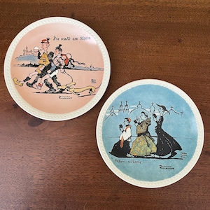 May include: Two round ceramic plates with raised beaded edges. The left plate has a peach background with the text Die walk am Rhein and an illustration of two figures and a dog. The right plate has a blue background with the text When in Rome and an illustration of three figures.