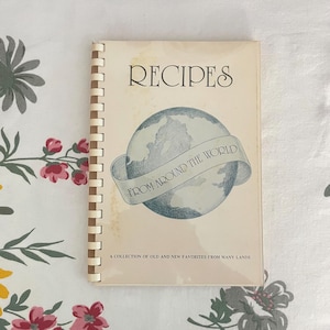 May include: A vintage cookbook titled "RECIPES FROM AROUND THE WORLD" with a spiral binding. The cover features a globe illustration with a banner. The text "A COLLECTION OF OLD AND NEW FAVORITES FROM MANY LANDS" is printed at the bottom.