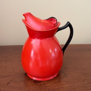 May include: A red ceramic pitcher with a black handle and a matching lid. The pitcher has a rounded body and a spout for pouring.