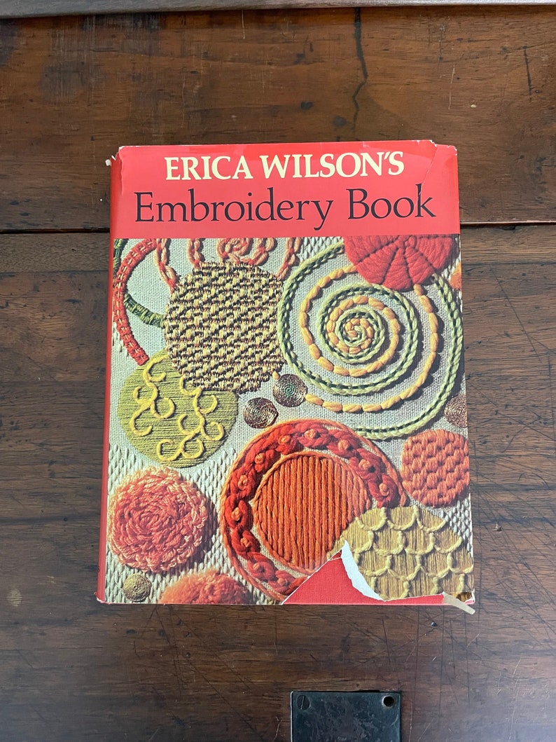 May include: A vintage copy of "Erica Wilson's Embroidery Book" with a red cover. The cover features colorful embroidered designs in orange, yellow, green, and brown. The book is on a dark wooden surface.