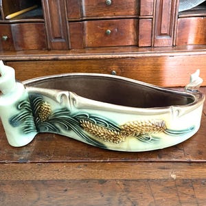 May include: A ceramic planter with a light green and brown color scheme. The planter features a curved design with decorative wheat and leaf patterns. The ends have stylized, upward-curving accents. The interior is a darker brown.