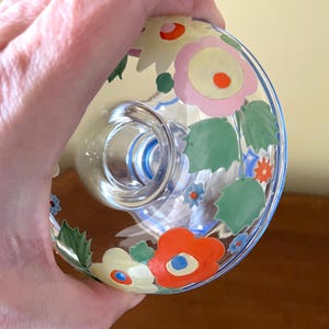 May include: A clear glass bottle stopper with a floral design. The stopper features a ring of flowers in pink, orange, and yellow with green leaves. The rim of the stopper is outlined in blue.