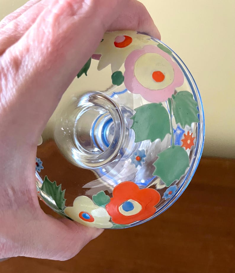May include: A clear glass bottle stopper with a floral design. The stopper features a ring of flowers in pink, orange, and yellow with green leaves. The rim of the stopper is outlined in blue.