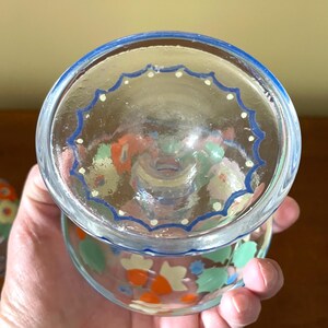 May include: Clear glass dessert cup with a blue rim and a blue and white scalloped design. The cup is decorated with colorful floral patterns in orange, green, and white. The cup is held in a hand.