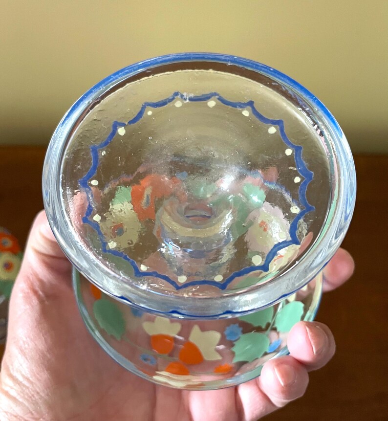 May include: Clear glass dessert cup with a blue rim and a blue and white scalloped design. The cup is decorated with colorful floral patterns in orange, green, and white. The cup is held in a hand.