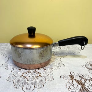 以下が含まれることがあります： 銅色の蓋とベース、銀色のボディのヴィンテージソースパン。ボディには花柄があります。黒いハンドルと蓋に黒いノブが付いています。ソースパンは白いレースのテーブルクロスの上にあります。