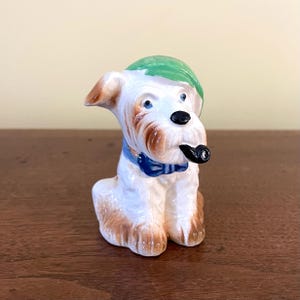 May include: A ceramic figurine of a seated dog with a green hat, blue bow tie, and a black pipe in its mouth. The dog has white fur with brown markings and blue eyes. The figurine is on a dark brown surface.