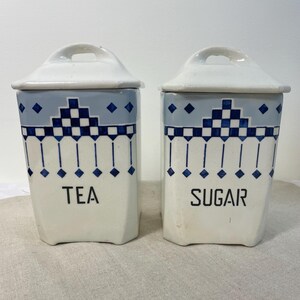 May include: Two white ceramic canisters with lids, labeled "TEA" and "SUGAR". Each canister has a blue and white geometric design around the top and a white handle on the lid. The canisters are on a neutral-colored surface.