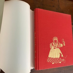 May include: A red hardcover book titled "Erica Wilson's Embroidery Book" with gold illustrations of a woman, dog, and bird. The book is open, revealing white pages. The cover has a textured appearance.