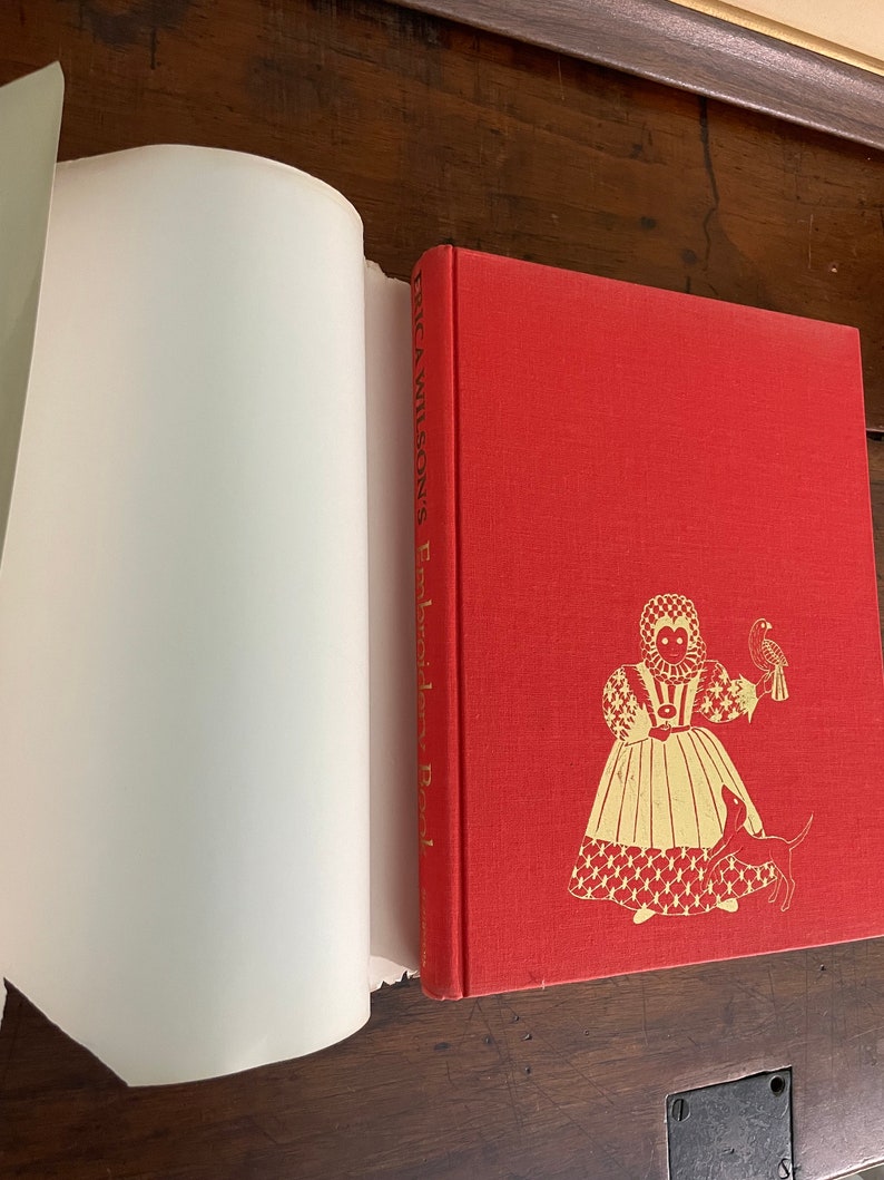 May include: A red hardcover book titled "Erica Wilson's Embroidery Book" with gold illustrations of a woman, dog, and bird. The book is open, revealing white pages. The cover has a textured appearance.