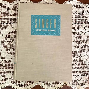 May include: A vintage Singer Sewing Book with a blue and white cover. The title "Singer Sewing Book" is printed in yellow on the cover.