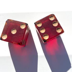 May include: Two vintage red dice with white dots, each showing different numbers. The dice are made of a translucent material, casting shadows on a white surface. The dice are a classic game accessory.