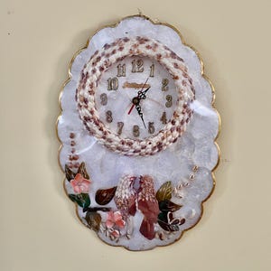May include: Oval wall clock with a white and gold scalloped frame. The clock face features a ring of seashells, a white background, and black numerals. Two birds are depicted at the bottom, with pink flowers and brown leaves.