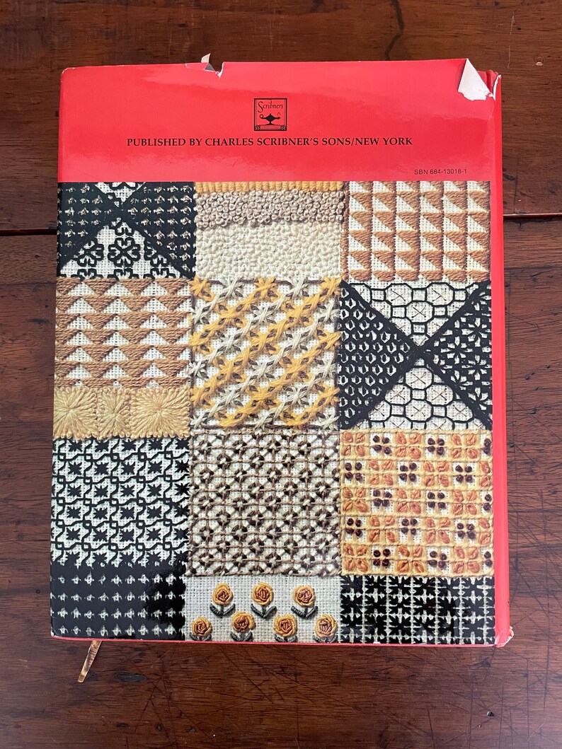 May include: A vintage book cover with a red top and a patchwork design of various needlepoint patterns in black, white, yellow, and brown. The text "PUBLISHED BY CHARLES SCRIBNER'S SONS/NEW YORK" is printed on the red top.