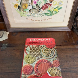 May include: An open book titled "ERICA WILSON'S Embroidery Book" rests on a wooden surface. The book's cover features colorful embroidery designs. In the background, a framed embroidery depicts a groundhog, rabbit, and deer among flowers.
