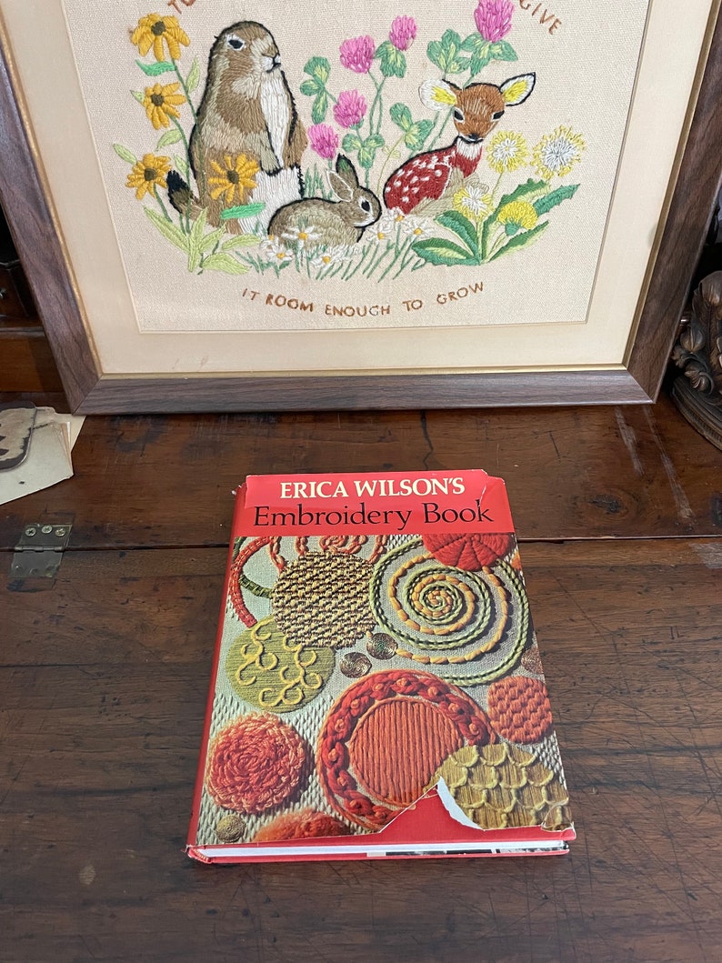 May include: An open book titled "ERICA WILSON'S Embroidery Book" rests on a wooden surface. The book's cover features colorful embroidery designs. In the background, a framed embroidery depicts a groundhog, rabbit, and deer among flowers.
