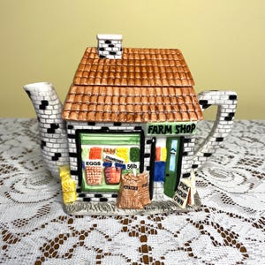May include: A ceramic teapot shaped like a farm shop. The teapot features a brown tiled roof, a white brick facade, and a green door. The shop window displays images of eggs, strawberries, and potatoes. The text "FARM SHOP" is above the window.