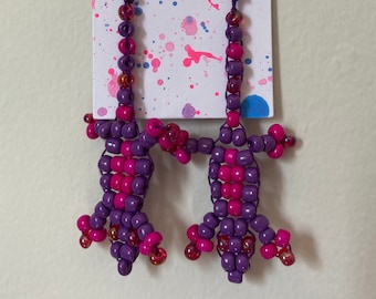 May include: A pair of purple and pink beaded earrings shaped like lizards. The earrings are hanging from a white card with the text "Spookie Jewelry" written in black.