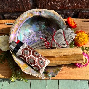 May include: A lighter encased in a beaded sleeve with a geometric honeycomb pattern in white, brown, and grey, resting inside an iridescent abalone shell alongside a bundle of dried sage tied with red and white string and a stick of palo santo wood.