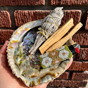 Peut inclure: Un coquillage ormeau irisé contenant un fagot de sauge blanche séchée lié par une ficelle jaune, deux bâtons de bois de palo santo brun clair et un briquet noir recouvert d'une gaine perlée à motifs floraux verts, blancs et dorés.