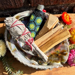 Puede incluir: Un cuenco de concha de abulón iridiscente que contiene un atado de salvia blanca envuelto en hilo rojo, dos palos de madera de palo santo de color marrón claro y un encendedor con funda de cuentas negra, verde y amarilla con patrón hexagonal.