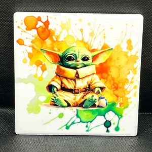 Puede incluir: Un personaje de dibujos animados verde y amarillo con orejas grandes, sentado sobre un fondo blanco con salpicaduras de acuarela de colores. El personaje lleva un mono marrón y amarillo con un cinturón marrón.