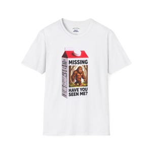 May include: A white short-sleeve t-shirt featuring a graphic of a milk carton with the text MISSING and HAVE YOU SEEN ME? printed on the front. The carton displays an illustration of a brown Bigfoot creature standing in a forest setting.