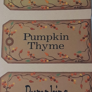 May include: Three rectangular, brown tags with black text and decorative borders. The tags read "Pumpkin Patch", "Pumpkin Thyme", and "Pumpkins". Each tag has a floral design with orange, yellow, and green accents.
