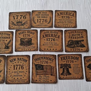 May include: A set of twelve square cork coasters featuring black printed designs related to 1776 and American history. Each coaster displays unique text and illustrations, including the Liberty Bell, an American flag, a cannon, and a tricorn hat.