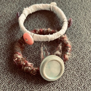May include: Two handmade bracelets. One is wrapped in white fabric and adorned with purple buttons. The other is wrapped in a multi-colored yarn and features a light blue button. Both are on a gray surface.