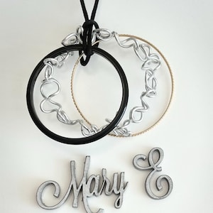 May include: A black and gold hoop pendant with silver wire detailing, suspended by a black cord. Below, the name "Mary E" is spelled out in silver-painted wooden letters. The design is set against a white background.