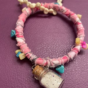 May include: A circular bracelet wrapped in pink and white plaid fabric, accented with silver wire, small turquoise-colored stones, and light-colored beads. A miniature glass bottle with a cork stopper containing white powder hangs from the bracelet.