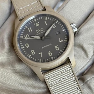 Peut inclure: Une montre-bracelet automatique IWC Schaffhausen avec un boîtier beige mat et un bracelet en tissu assorti. Le cadran gris foncé présente des chiffres arabes blancs, un guichet de date à trois heures et un triangle à douze heures.