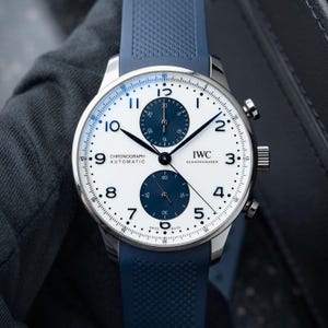 Peut inclure: Une montre chronographe automatique IWC Schaffhausen avec un cadran blanc, deux sous-cadrans bleu foncé et des chiffres arabes noirs. La montre présente un boîtier en acier inoxydable argenté et un bracelet en caoutchouc bleu texturé.