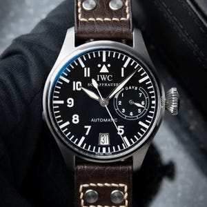 Peut inclure: Une montre-bracelet automatique IWC Schaffhausen avec un cadran noir, des chiffres arabes blancs, un guichet de date à six heures et un indicateur de réserve de marche. La montre possède un boîtier argenté et un bracelet en cuir marron à rivets.