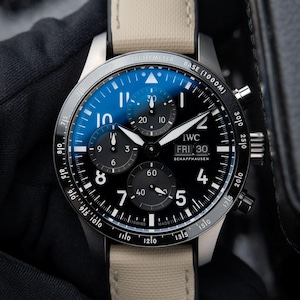 Peut inclure: Une montre chronographe IWC Schaffhausen avec un cadran noir, des chiffres arabes blancs et trois sous-cadrans. La montre présente une lunette tachymètre, un guichet jour-date à trois heures et un bracelet en tissu beige avec surpiqûres blanches.