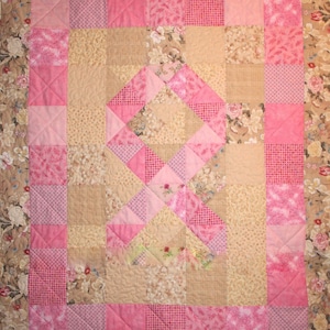 May include: A pink and beige quilt with a pink ribbon design in the center. The quilt is made of various fabrics with floral patterns and different shades of pink.