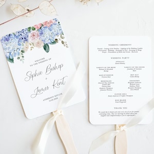 May include: A white wedding program fan with a watercolor floral design of blue and pink flowers. The fan has a white ribbon tied around the handle. The text on the fan reads "Welcome to the Wedding of Sophie Bishop + James Kent, Anderson Estate, 12.05.2025".