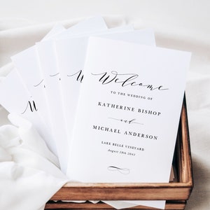 Op de afbeelding: Welkom op de bruiloft van Katherine Bishop en Michael Anderson in Lake Belle Vineyard op 20 augustus 2045. Een stapel witte huwelijksboekjes met zwarte kalligrafie.