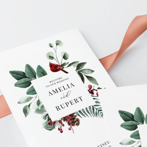 May include: Wedding invitation featuring a watercolor design with green leaves, red berries, and a red cardinal. The invitation reads "Welcome to our Wedding" and the names "Amelia and Rupert". A peach ribbon is in the background.