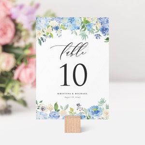 Pode incluir: Cartão de número de mesa de casamento floral azul e branco com o número 10 e o texto "mesa" em uma fonte cursiva. O cartão também inclui os nomes "Kristina & Michael" e a data "18 de agosto de 2045".