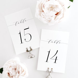 Pode incluir: Dois números de mesa de cartão branco com caligrafia preta. Os números são 14 e 15. Os cartões são mantidos por suportes de números de mesa prateados. O fundo é branco.