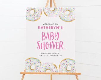 Puede incluir: Bienvenido al Baby Shower de Katheryn con temática de donas. El letrero es blanco con texto rosa y está decorado con tres donas rosadas glaseadas con chispas de colores.