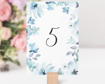 May include: A table number card with the number 5 in black script, surrounded by a watercolor floral design in shades of blue and green. The card is held by a small wooden block, perfect for weddings or events.